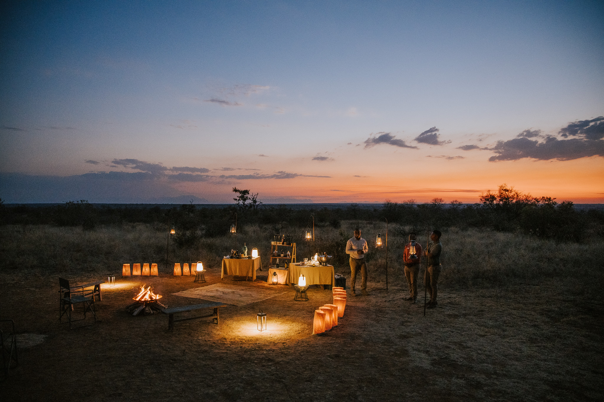 Lantern-lit outdoor dining setup in the African bush at sunset.