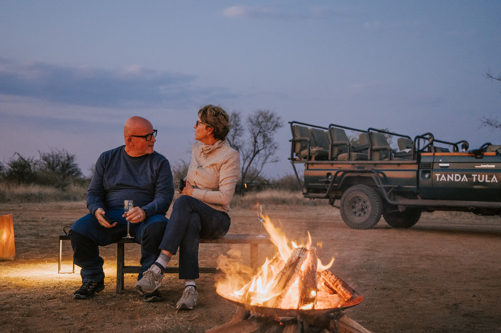 Couple enjoying sundowners by a fire on safari with game vehicle nearby.