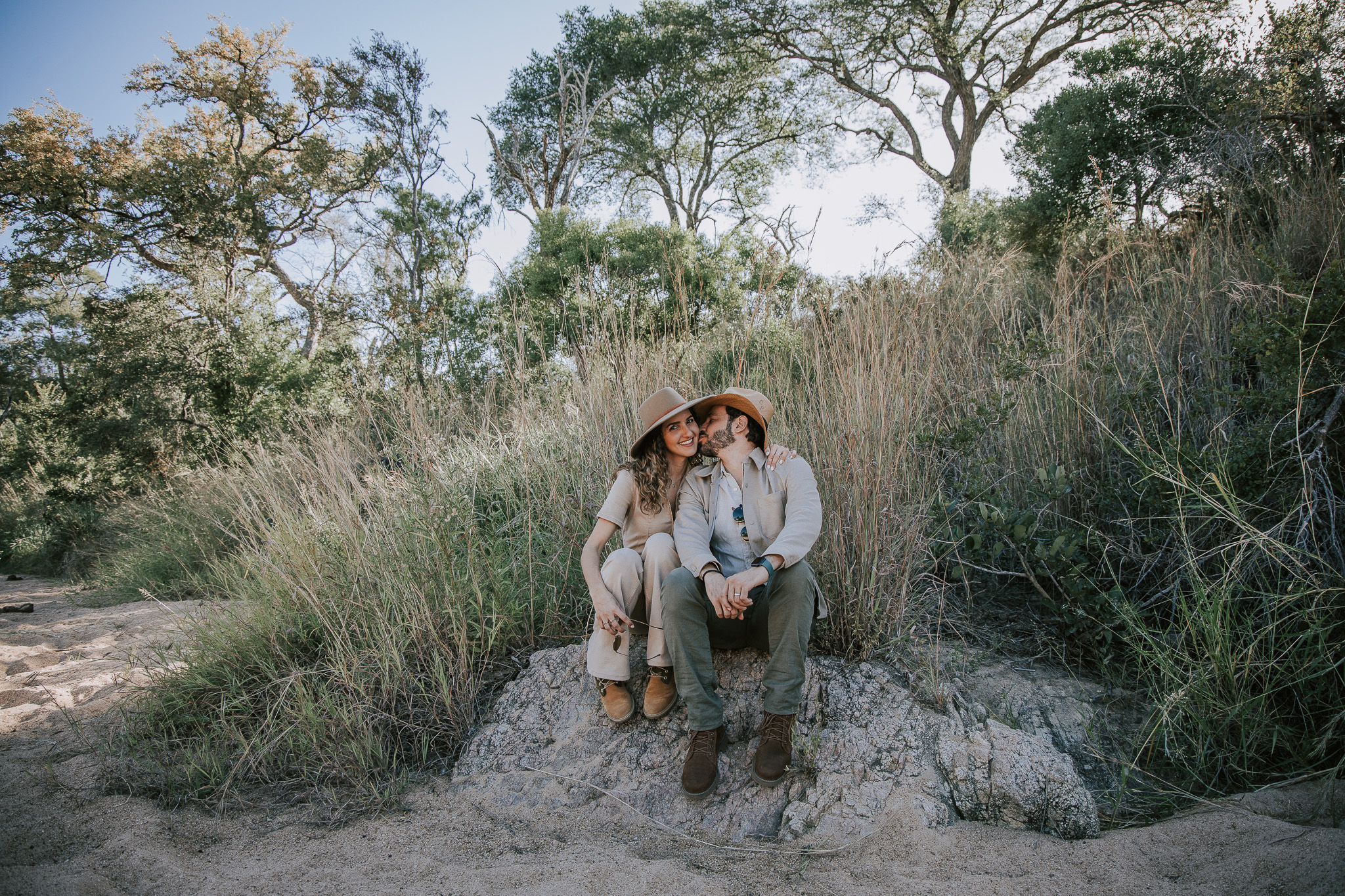 Couple sitting together outdoors, wearing safari hats and sharing a romantic moment.