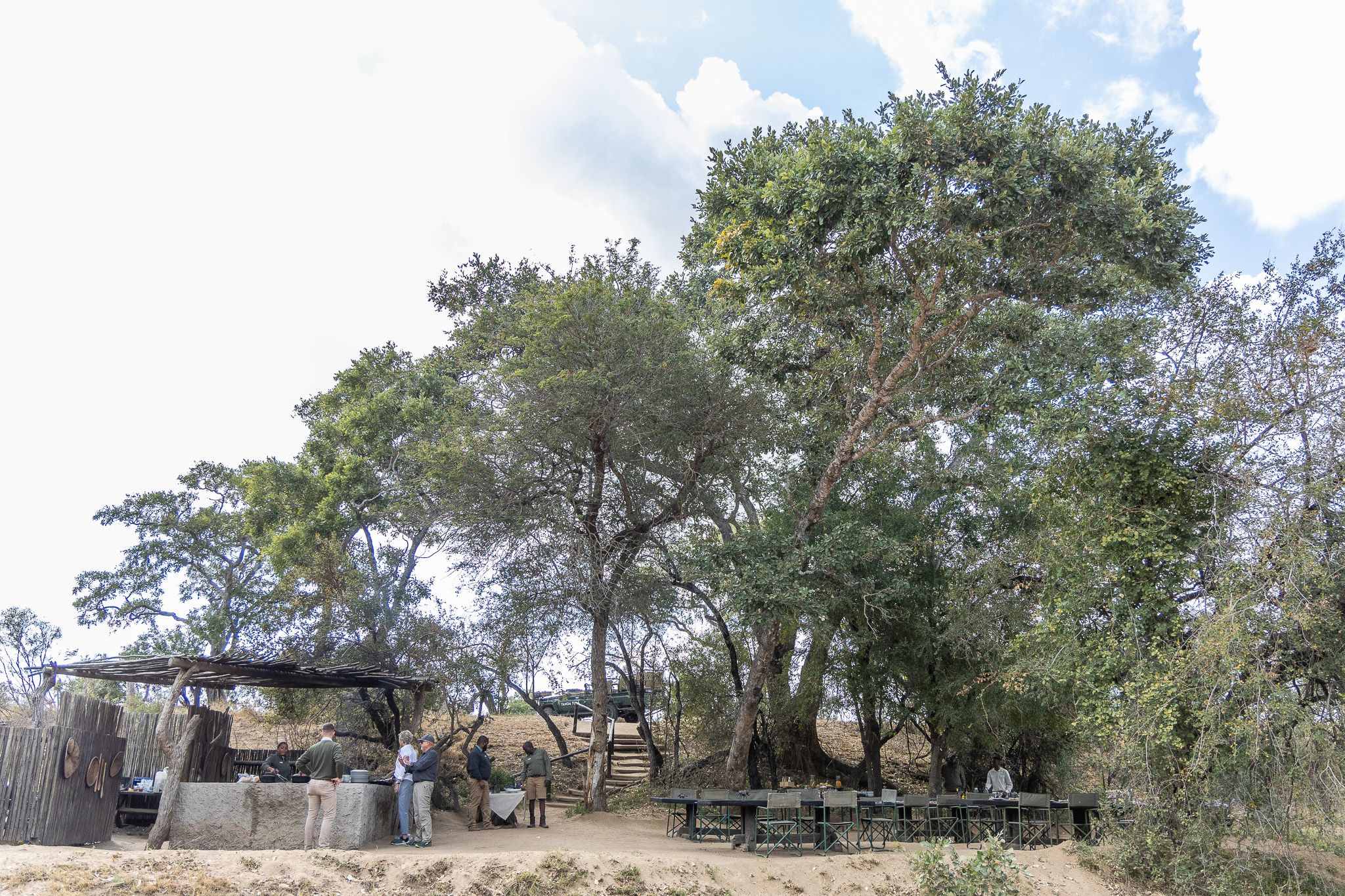 A wide view of an outdoor bush breakfast setting at Tanda Tula, with staff preparing food under a rustic thatched canopy, and long communal tables set beneath the trees in the Timbavati.