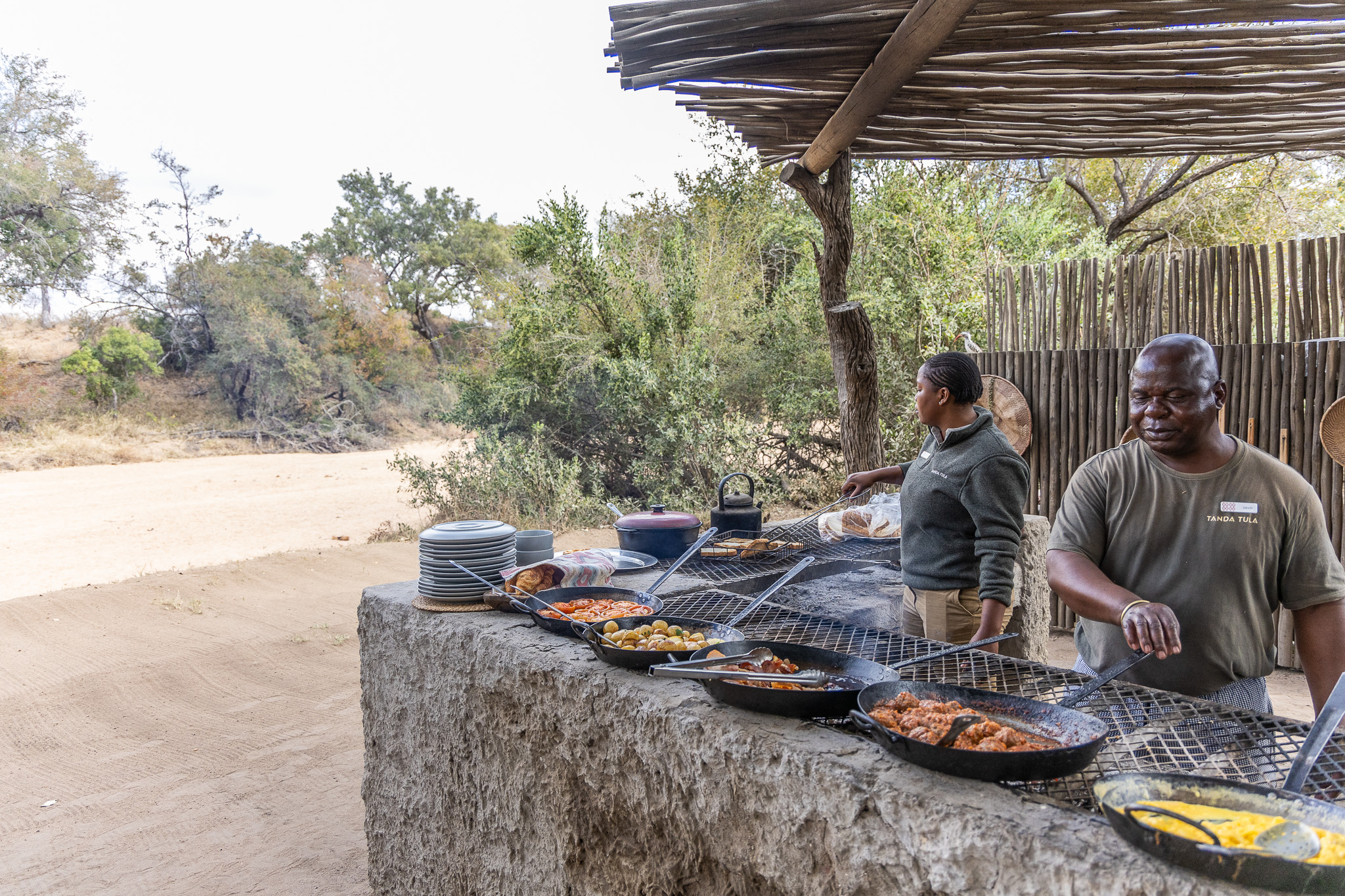 Tanda Tula team members preparing a bush breakfast over open flames in the Timbavati wilderness, with an array of cast iron pans filled with vibrant, freshly cooked dishes.