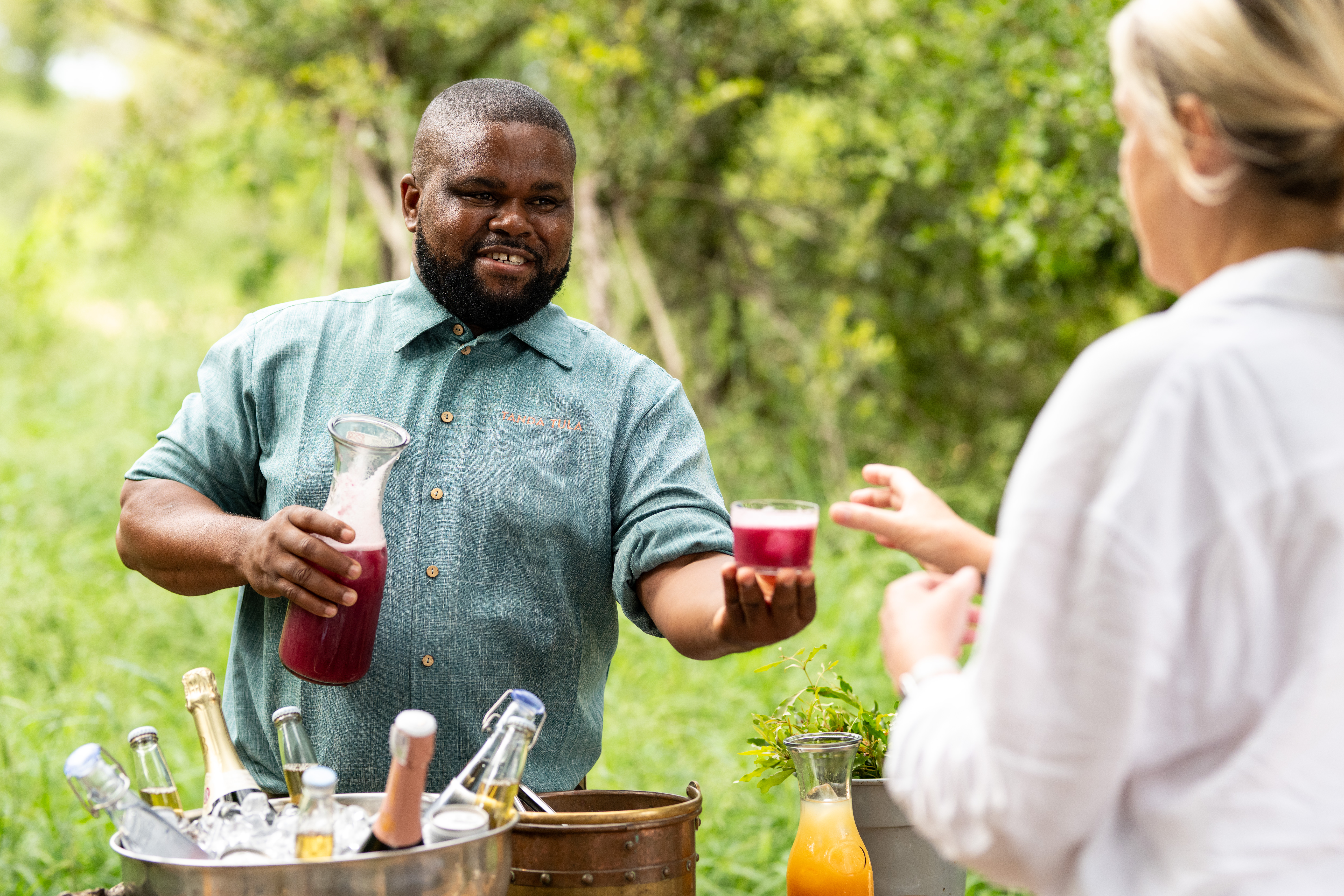 A smiling Tanda Tula team member offers a glass of fresh beetroot juice to a guest during an outdoor drinks service, surrounded by lush Timbavati greenery and a selection of chilled beverages.