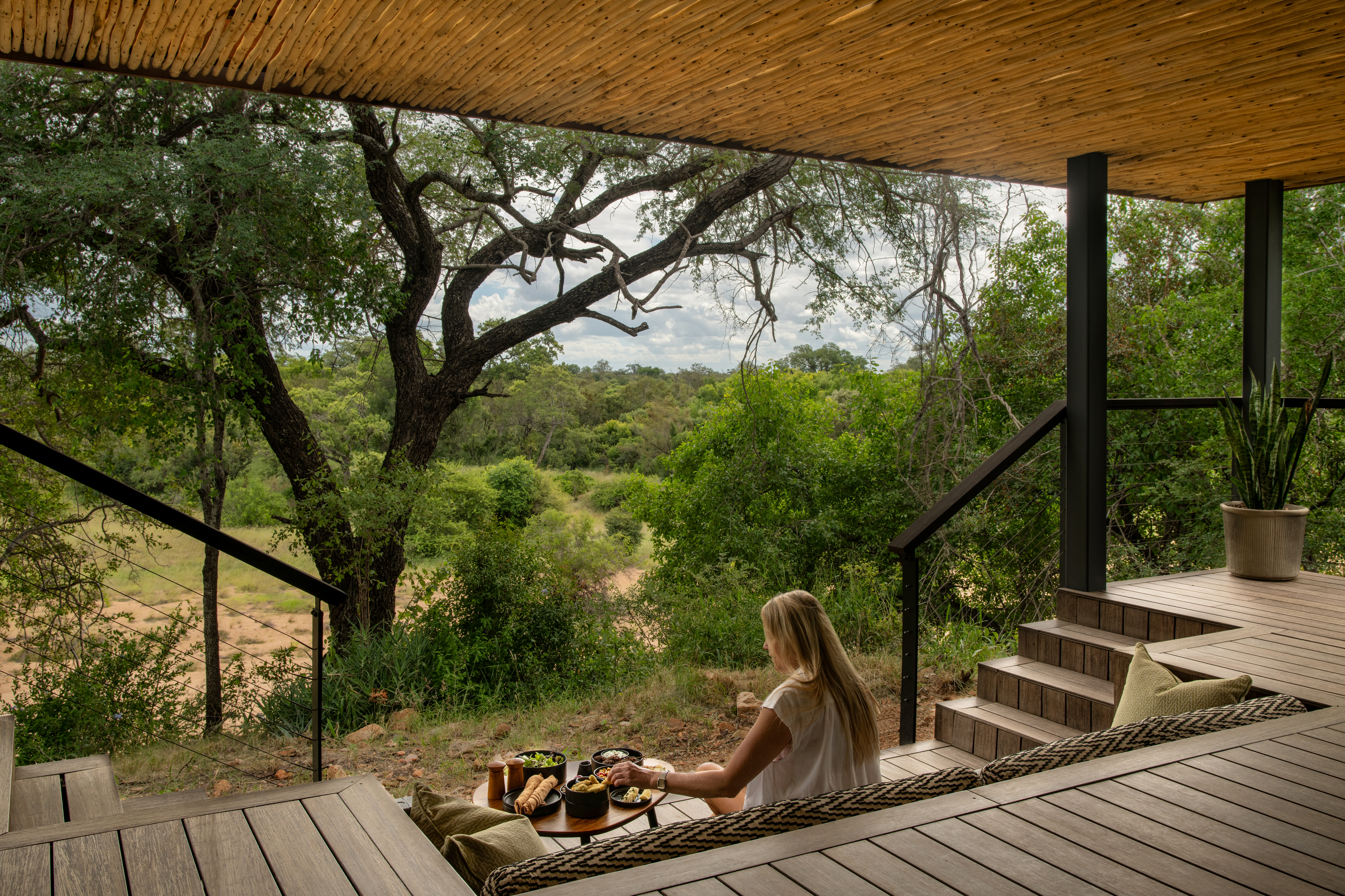 A woman enjoys a private lunch on the shaded deck of her luxury safari suite at Tanda Tula, overlooking the lush green Timbavati riverbed and surrounding wilderness.