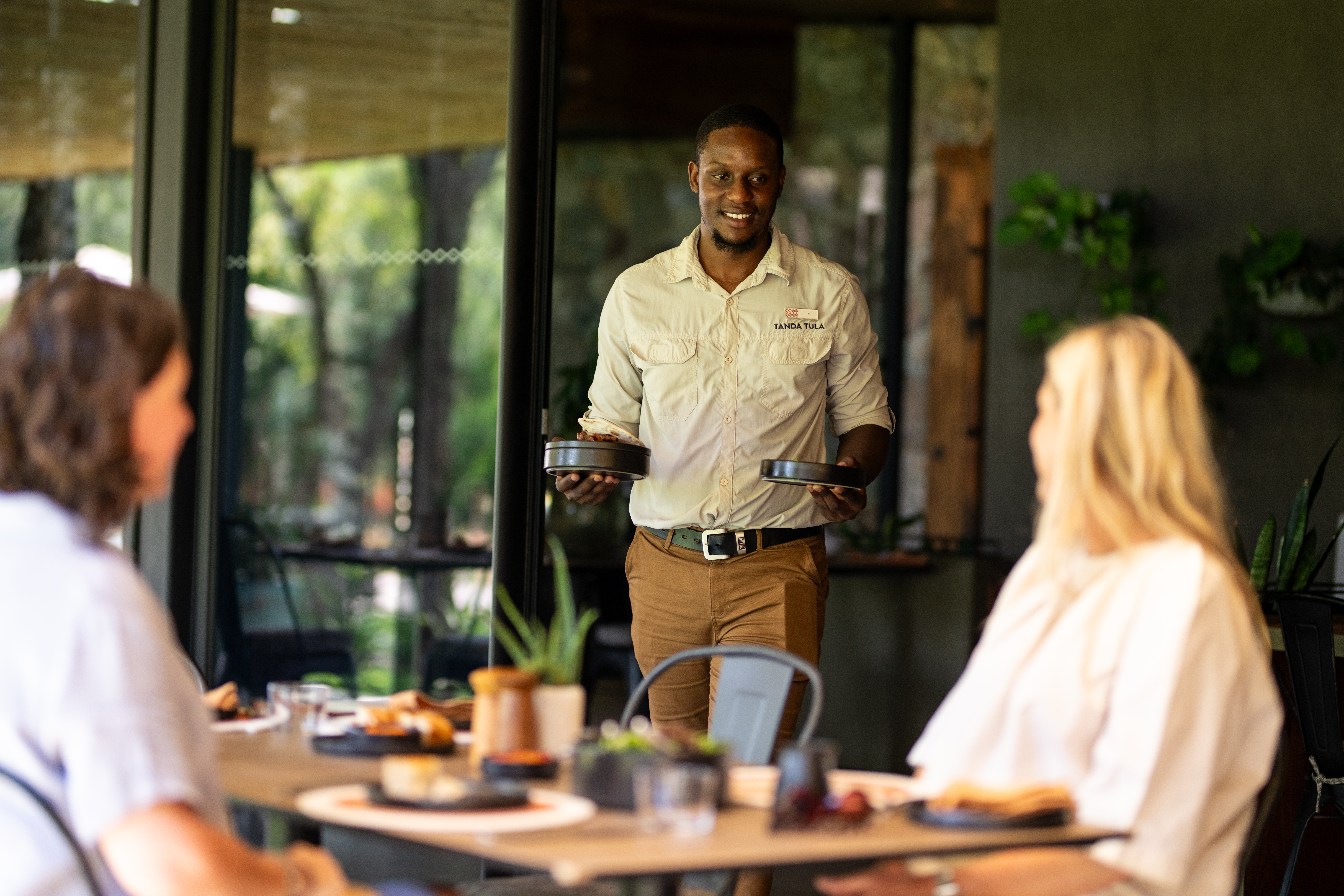 A smiling Tanda Tula staff member serves freshly prepared lunch to two guests on the outdoor patio, capturing the warmth, connection, and personalised service that define the lodge’s soulful hospitality experience.
