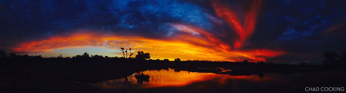 Panoramic iPhone photo of a dramatic sunset over a waterhole with vivid reds, oranges, and deep blue clouds.