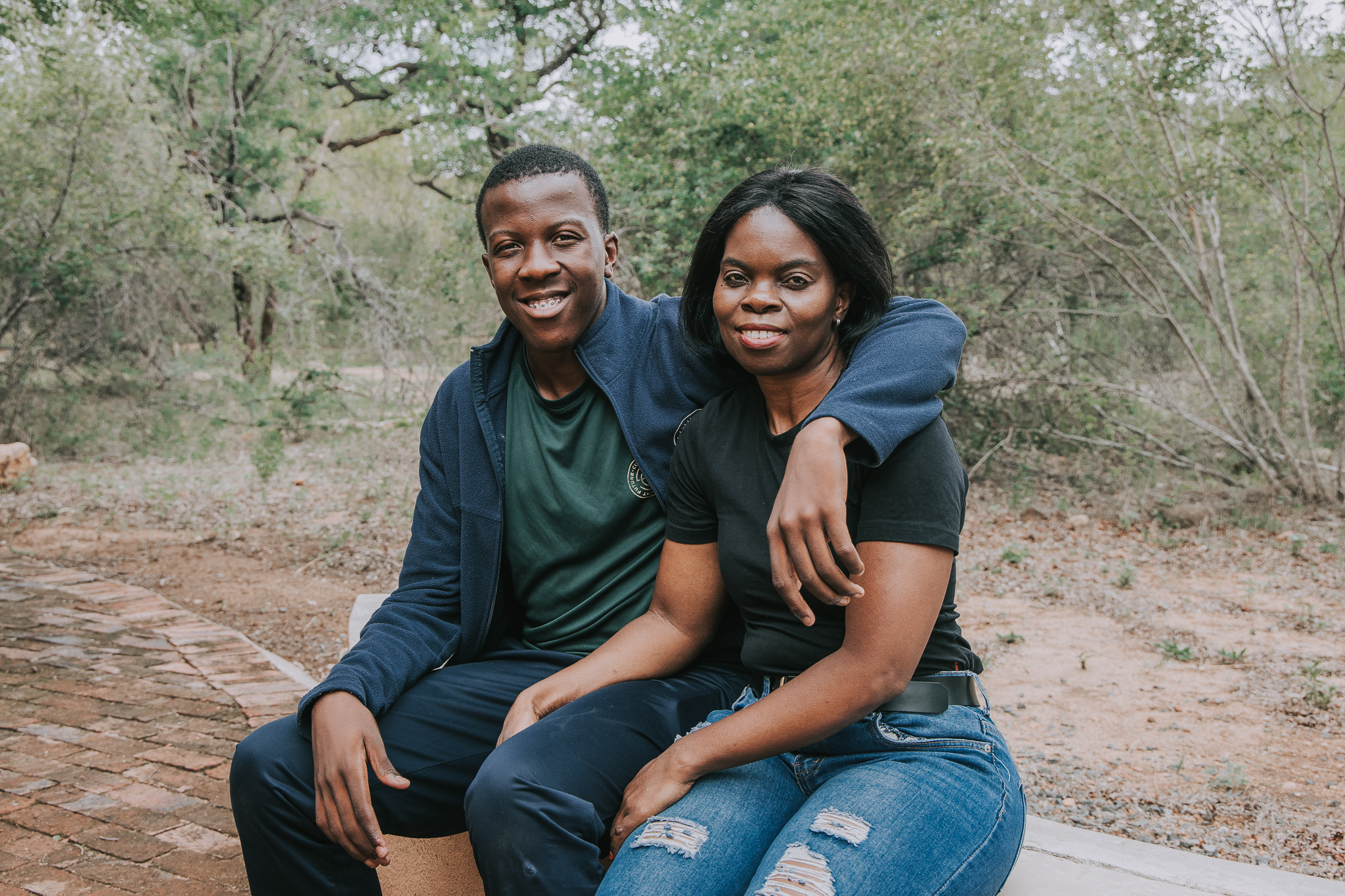 A young man and a woman sitting outdoors with his arm around her shoulder, both smiling.