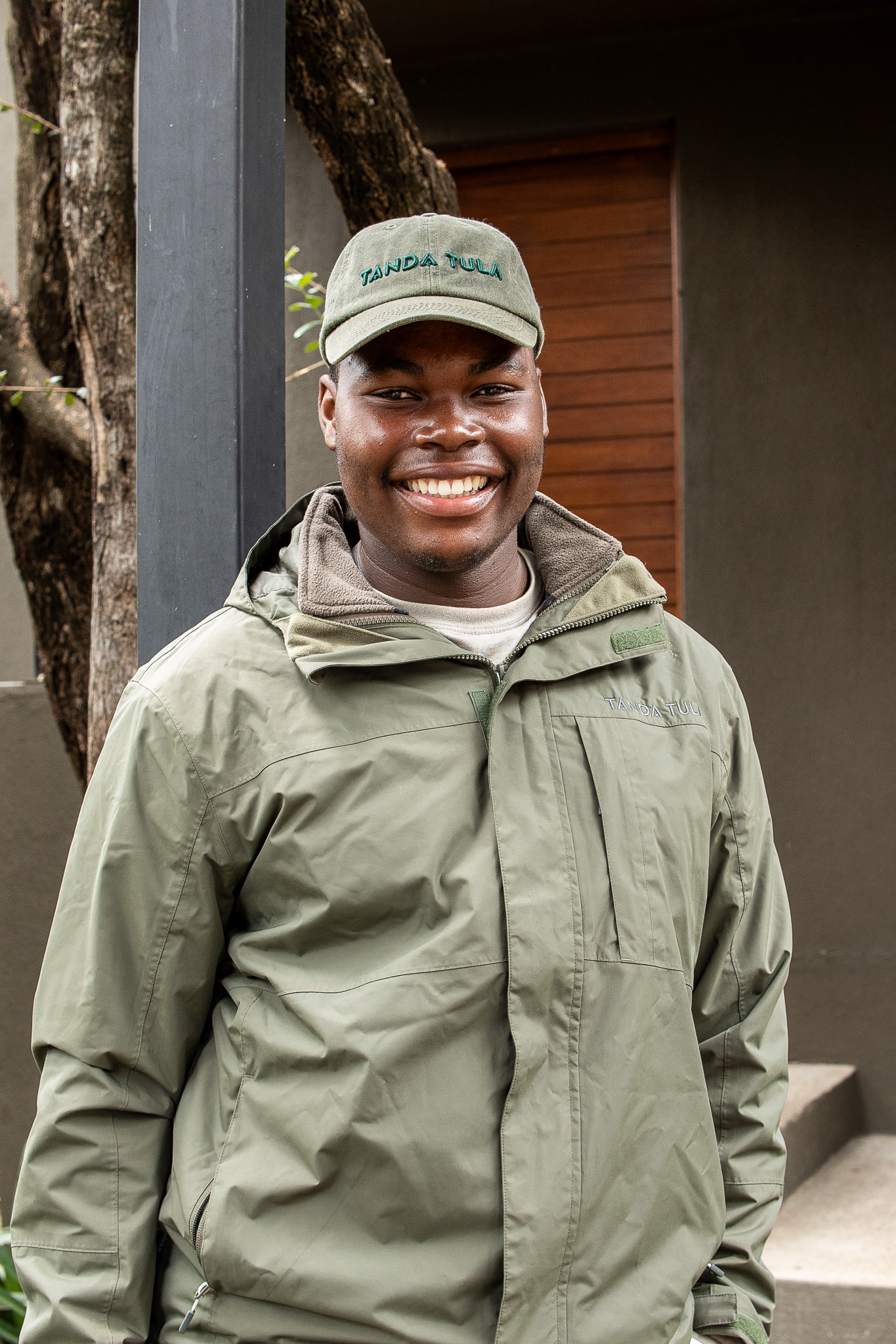 A young Tanda Tula team member wearing an olive jacket and cap, smiling outdoors.