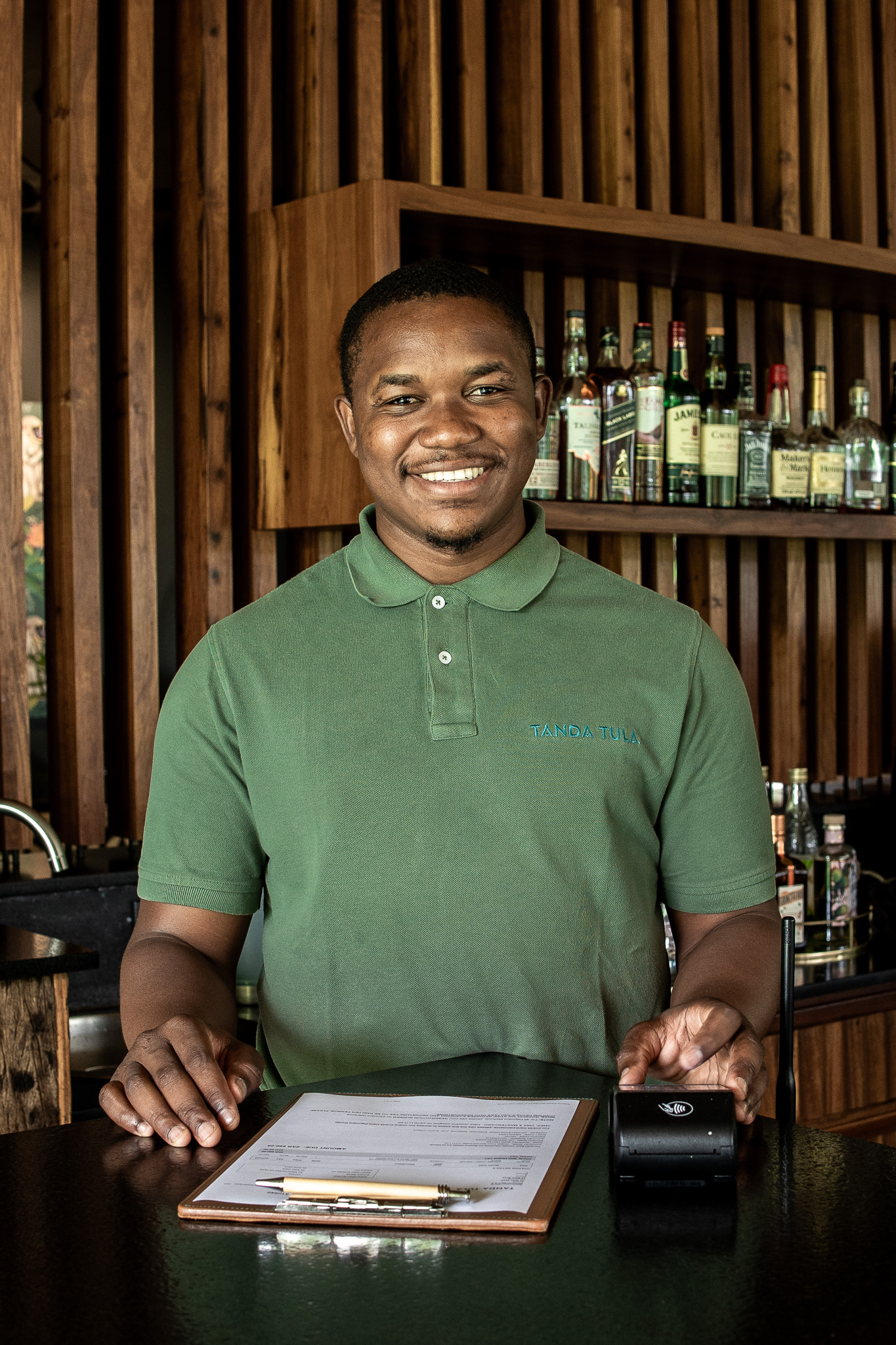 A Tanda Tula team member standing behind the bar counter, smiling with a clipboard and card machine in front of him.