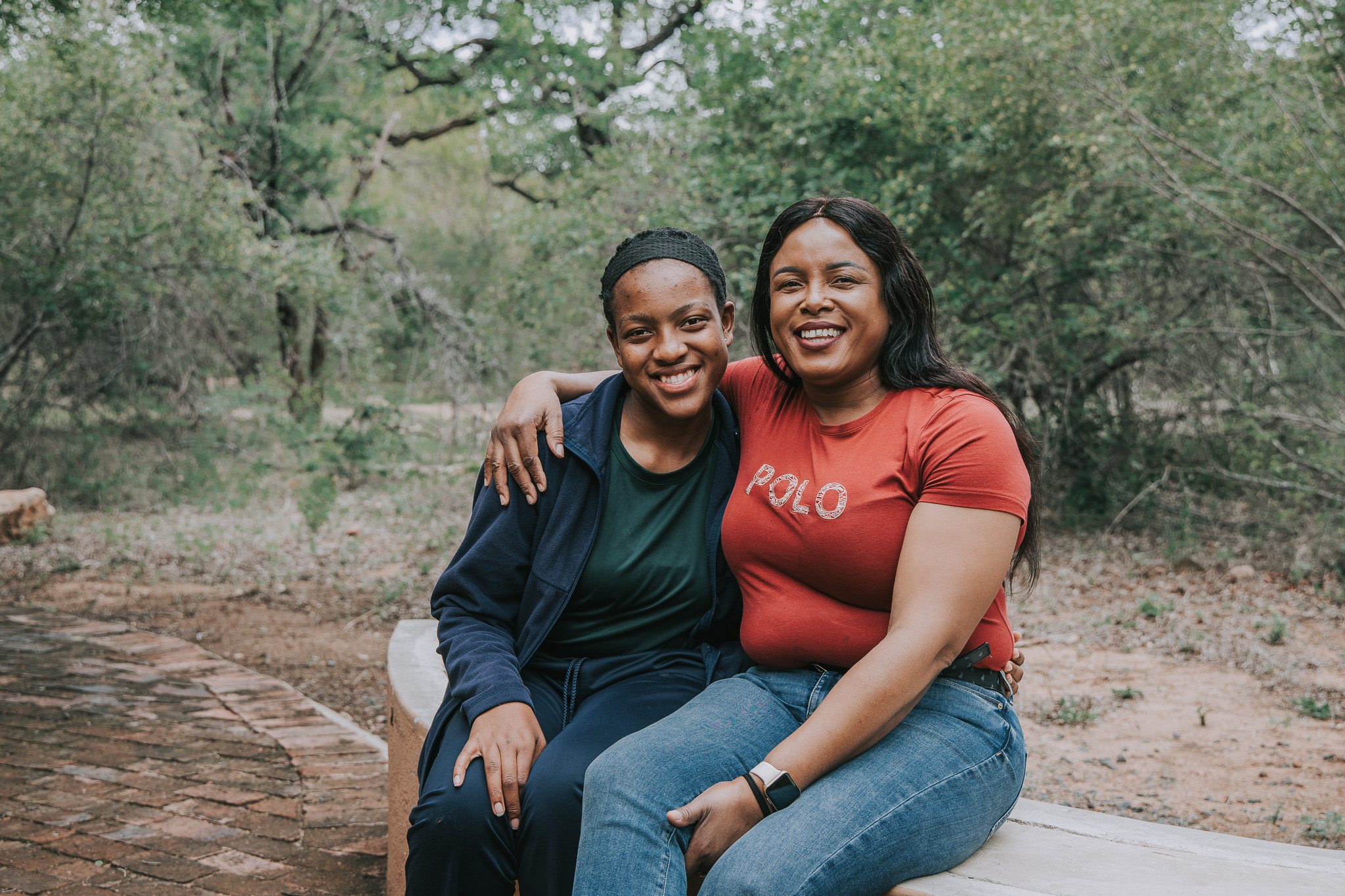 Two women sitting outdoors with arms around each other, smiling warmly at the camera.